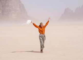 Sotto le Stelle dell’Austria / Kulturforum Rom Das Bild zeigt Schauspielerin Vicky Krieps in ihrer Rolle als Ingeborg Bachmann im Film „Reise in die Wüste“. Sie geht über hellen Wüstensand. Sie trägt eine orangefarbene Bluse, eine beige Hose und ein buntes Kopftuch. In der linken Hand hält sie einen weißen Schal, der im Wind weht. Lachend wirft sie die Arme nach oben. Im Hintergrund sind Felsen zu sehen.