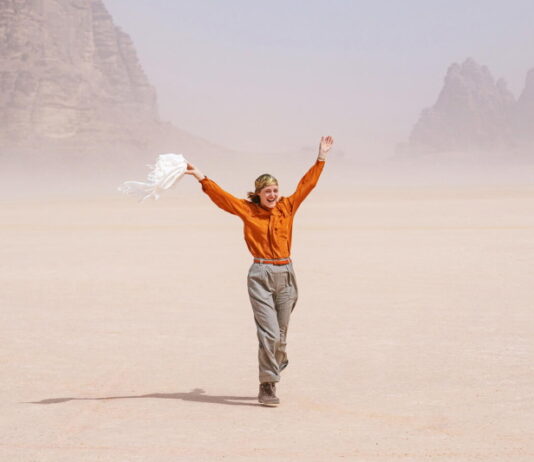 Sotto le Stelle dell’Austria / Kulturforum Rom Das Bild zeigt Schauspielerin Vicky Krieps in ihrer Rolle als Ingeborg Bachmann im Film „Reise in die Wüste“. Sie geht über hellen Wüstensand. Sie trägt eine orangefarbene Bluse, eine beige Hose und ein buntes Kopftuch. In der linken Hand hält sie einen weißen Schal, der im Wind weht. Lachend wirft sie die Arme nach oben. Im Hintergrund sind Felsen zu sehen.