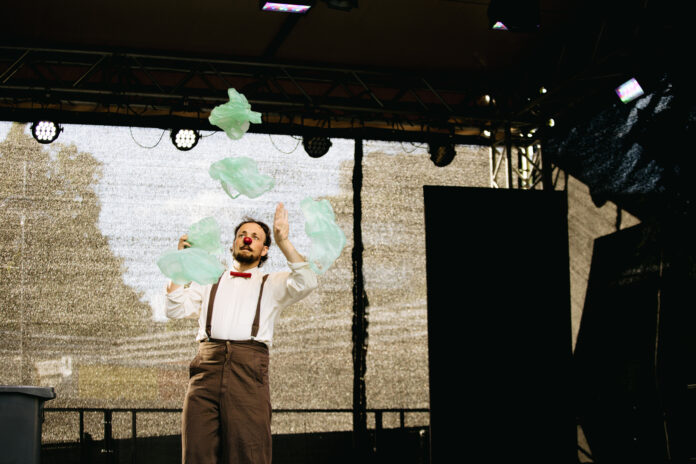 Das Bild zeigt ein melancholisch blickender Clown mit roter Nase jongliert mit Plastiktüten auf einer Open-Air Bühne
