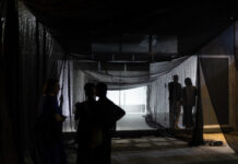 At the Interstice / Kulturforum Accra Das Bild zeigt einen Ausstellungsraum mit einer weißen Wand. Links im Bild steht eine Person im Profil und betrachtet eine großflächige Installation an der Wand. Die Person trägt eine dunkle Jacke und eine dunkle Hose und hat eine Glatze. Die Installation besteht aus zahlreichen kleinen, überwiegend schwarz-weißen Fotografien und Bildausschnitten. Sie sind in unregelmäßigen Abständen auf der weißen Wand angeordnet und bilden gemeinsam eine größere, offene Form. Im Vordergrund sind unscharfe helle und dunkle Formen zu sehen. Im Hintergrund links sind große Fenster oder Wandöffnungen des Ausstellungsraums erkennbar. Die Aufnahme ist insgesamt in gedämpften Grau-, Schwarz- und Weißtönen gehalten.