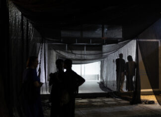 At the Interstice / Kulturforum Accra Das Bild zeigt einen Ausstellungsraum mit einer weißen Wand. Links im Bild steht eine Person im Profil und betrachtet eine großflächige Installation an der Wand. Die Person trägt eine dunkle Jacke und eine dunkle Hose und hat eine Glatze. Die Installation besteht aus zahlreichen kleinen, überwiegend schwarz-weißen Fotografien und Bildausschnitten. Sie sind in unregelmäßigen Abständen auf der weißen Wand angeordnet und bilden gemeinsam eine größere, offene Form. Im Vordergrund sind unscharfe helle und dunkle Formen zu sehen. Im Hintergrund links sind große Fenster oder Wandöffnungen des Ausstellungsraums erkennbar. Die Aufnahme ist insgesamt in gedämpften Grau-, Schwarz- und Weißtönen gehalten.