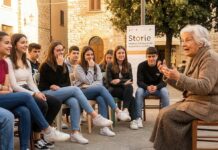 STORIE / Kulturforum Rom Das Bild zeigt eine ältere Dame, die Jugendlichen etwas erzählt. Die Gruppe sitzt im Halbkreis und folgt der Dame amüsiert und gespannt. Es ist eine gute Stimmung. Von der Architektur erkennt man die italienische Dorfschaft, im Hintergrund ist das Plakat vom Festival zu sehen.