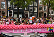 World Pride / Botschaft Den Haag Das Bild zeigt feiernde Menschen an den Amsterdamer Grachten mit Regenbogenfahnen sowie ein Banner mit der Aufschrift „United in Pride“.