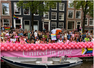 World Pride / Botschaft Den Haag Das Bild zeigt feiernde Menschen an den Amsterdamer Grachten mit Regenbogenfahnen sowie ein Banner mit der Aufschrift „United in Pride“.