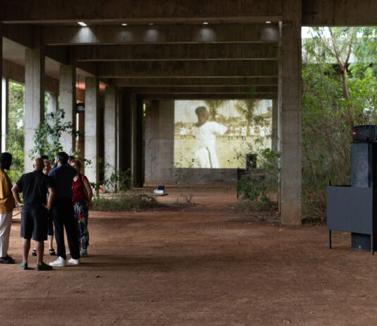 Speakers / Kulturforum Accra Unter einem offenen, verwitterten Betonbau steht eine Gruppe von fünf Personen auf einem erdigen Boden. Im Hintergrund ist auf einer großen Projektionsfläche die unscharfe Aufnahme einer schwarzen Person in weißer Kleidung zu sehen. Rechts stehen mehrere Lautsprecher, deren Anordnung an eine Skulptur erinnert. Sie sind von einem schwarzen, quaderförmigen Rahmen eingefasst. Durch die offenen Seiten des Gebäudes ist dichtes grünes Buschwerk sichtbar. Die Szene wirkt wie eine Kunstinstallation oder eine Filmvorführung im Freien.