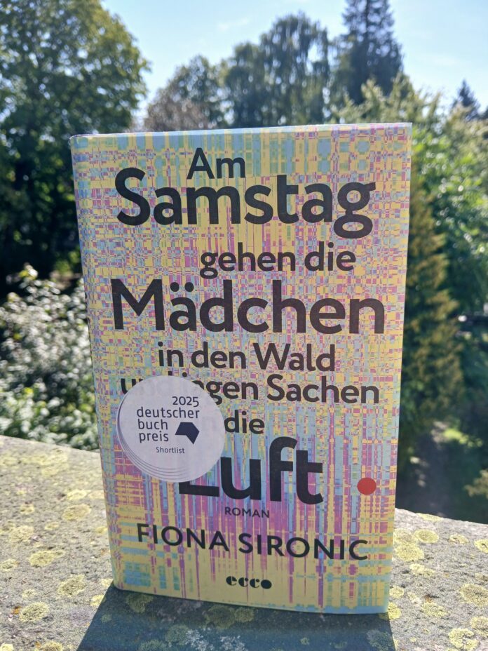 Auf einem steinernen Mauersims steht im Freien das Buch Am Samstag gehen die Mädchen in den Wald und jagen Sachen in die Luft von Fiona Sironic. Das Cover ist in Gelb, Rosa, Blau und Violett gestaltet und zeigt ein pixelartiges, abstraktes Muster. Auf dem Cover steht außerdem „2025 deutscher buchpreis Shortlist“. Im Hintergrund sind unscharf grüne Bäume und blauer Himmel zu sehen.
