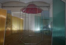 Ausstellung Darja Shatalova „Metonymia“ / Kulturforum Moskau Das Bild zeigt eine Installation aus Metall-Konstruktionen mit Plastik, zentraler Teil hat ein bemaltes Dach mit Zeichnungen in rot, links und rechts von diesem so genannten stehenden Turm sind die Wände mit einem durchsichtigen Plastik, links bemalt mit Zeichnungen in gelb, rechts bemalt mit Zeichnungen in blau. Die ganze Installation steht in einem leeren Raum, eine Zeche der ehemaligen Papierfabrik, zwei Ausschnitte der hinteren Wand links und rechts sind mit den blauen Fliesen ausgelegt. Vom hinten vom Boden wird die Installation noch beleuchtet.