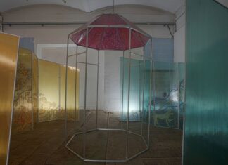Ausstellung Darja Shatalova „Metonymia“ / Kulturforum Moskau Das Bild zeigt eine Installation aus Metall-Konstruktionen mit Plastik, zentraler Teil hat ein bemaltes Dach mit Zeichnungen in rot, links und rechts von diesem so genannten stehenden Turm sind die Wände mit einem durchsichtigen Plastik, links bemalt mit Zeichnungen in gelb, rechts bemalt mit Zeichnungen in blau. Die ganze Installation steht in einem leeren Raum, eine Zeche der ehemaligen Papierfabrik, zwei Ausschnitte der hinteren Wand links und rechts sind mit den blauen Fliesen ausgelegt. Vom hinten vom Boden wird die Installation noch beleuchtet.