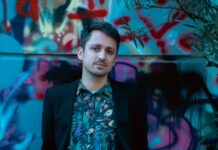 Literatur in Göteborg und Stockholm / Botschaft Stockholm Das Bild zeigt den Autor Fabian Navarro in einem schwarzen Sakko und einem bunten Hemd mit Vogelmuster. Im Hintergrund ist eine ebenso bunte, mit Graffiti bemalte, Wand zu sehen