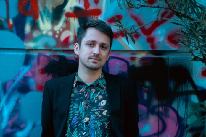 Das Bild zeigt den Autor Fabian Navarro in einem schwarzen Sakko und einem bunten Hemd mit Vogelmuster. Im Hintergrund ist eine ebenso bunte, mit Graffiti bemalte, Wand zu sehen