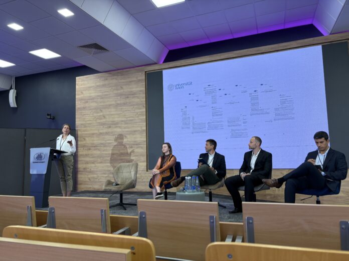 Foto eines Podiumsgesprächs an der Kozminski University. Prof. Iris Eisenberger spricht am Rednerpult, während vier weitere Teilnehmerinnen und Teilnehmer auf der Bühne daneben sitzen. Im Hintergrund ist eine Präsentation der Universität Wien zum Thema „Guidelines and Code of Practice on Trans¬parent AI“ zu sehen.
