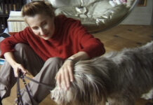 Die Rolle des Körpers im Werk Elfriede Jelineks / Kulturforum Madrid Das Bild zeigt Elfride Jelinek in einem roten Pullover und grauer Hose gekleidet, mit einem Hund an der Leine am Boden sitzend, den sie streichelt.