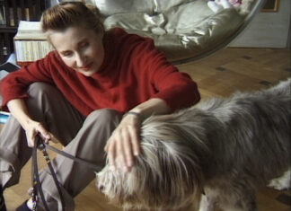 Die Rolle des Körpers im Werk Elfriede Jelineks / Kulturforum Madrid Das Bild zeigt Elfride Jelinek in einem roten Pullover und grauer Hose gekleidet, mit einem Hund an der Leine am Boden sitzend, den sie streichelt.