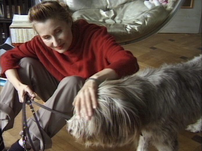 Das Bild zeigt Elfride Jelinek in einem roten Pullover und grauer Hose gekleidet, mit einem Hund an der Leine am Boden sitzend, den sie streichelt.