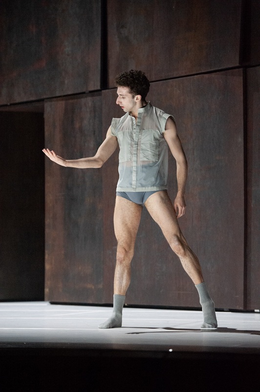 Das Bild zeigt den Tänzer Gabriele Aime in einem halbtransparenten, ärmellosen Oberteil und grau-blauer Unterwäsche auf einer Bühne in Tanzpose vor dunkler, rostbrauner Kulisse und blickt auf seine geöffnete Hand.