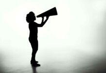 Stimmen. Spuren und Séancen / Kulturforum Berlin Das Bild zeigt den dunklen Umriss einer Person mit längeren Haaren, die in ein Megafon schreit. Der Hintergrund ist verschwommen weiß und grau.
