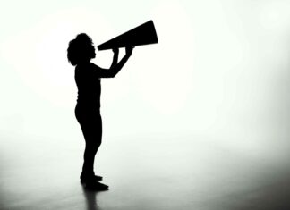 Stimmen. Spuren und Séancen / Kulturforum Berlin Das Bild zeigt den dunklen Umriss einer Person mit längeren Haaren, die in ein Megafon schreit. Der Hintergrund ist verschwommen weiß und grau.