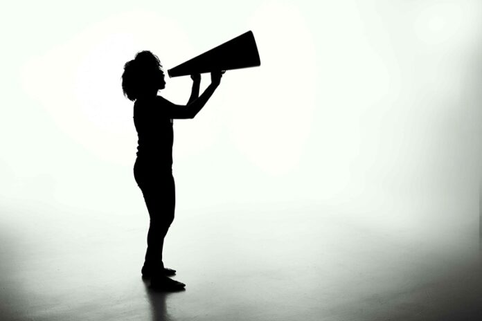 Foto: Patrick Fore / Unsplash Das Bild zeigt den dunklen Umriss einer Person mit längeren Haaren, die in ein Megafon schreit. Der Hintergrund ist verschwommen weiß und grau.