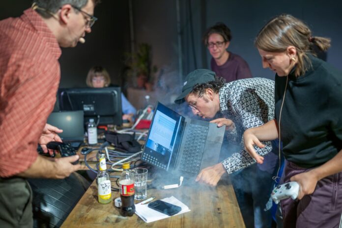 Das Foto zeigt eine Szene aus der Performance, man sieht eine Gruppe von Leuten um einen rauchenden Laptop versammelt.