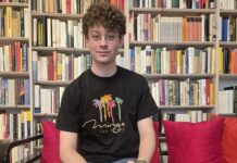 Ezequiel Max: Das Ende des Schweigens / wissensART Ein junger Mann sitzt auf einer Couch und blickt gerade in die Kamera, dahinter ein Bücherregal mit vielen Büchern.