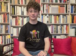 Ezequiel Max: Das Ende des Schweigens / wissensART Ein junger Mann sitzt auf einer Couch und blickt gerade in die Kamera, dahinter ein Bücherregal mit vielen Büchern.