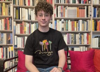 Ezequiel Max: Das Ende des Schweigens / Podcast wissensART Ein junger Mann sitzt auf einer Couch und blickt gerade in die Kamera, dahinter ein Bücherregal mit vielen Büchern.