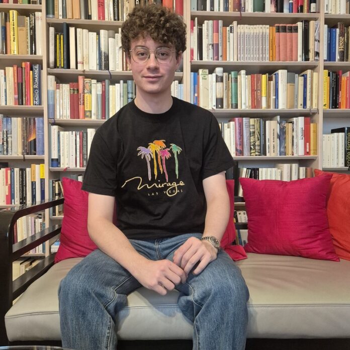 Bild: Elisabeth J. Nöstlinger Ein junger Mann sitzt auf einer Couch und blickt gerade in die Kamera, dahinter ein Bücherregal mit vielen Büchern.