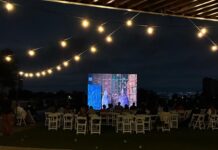 EU-Filmfestival Ghana / Kulturforum Accra Das Bild zeigt eine abendliche Open-Air-Kinovorführung auf einer Dachterrasse. Das Publikum sitzt auf weißen Klappstühlen vor einer großen Leinwand, auf der ein Film läuft; darüber spannen sich warm leuchtende Lichterketten.