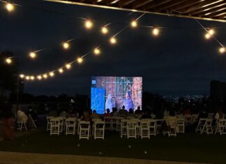 EU-Filmfestival Ghana / Kulturforum Accra Das Bild zeigt eine abendliche Open-Air-Kinovorführung auf einer Dachterrasse. Das Publikum sitzt auf weißen Klappstühlen vor einer großen Leinwand, auf der ein Film läuft; darüber spannen sich warm leuchtende Lichterketten.