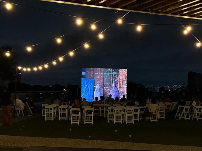 Foto: Kulturforum Accra Das Bild zeigt eine abendliche Open-Air-Kinovorführung auf einer Dachterrasse. Das Publikum sitzt auf weißen Klappstühlen vor einer großen Leinwand, auf der ein Film läuft; darüber spannen sich warm leuchtende Lichterketten.